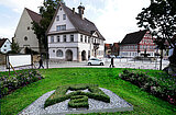 Blick auf einen grünen Platz mit einem Heckenornament in Form eines Wappens, umgeben von Blumenbeeten, davor historische Fachwerkhäuser und ein weißes Auto auf der Straße