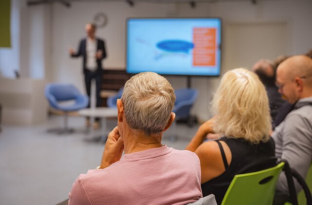 Rückansicht von drei sitzenden Personen, die einem stehenden Vortragenden mit Präsentation auf einem Bildschirm folgen