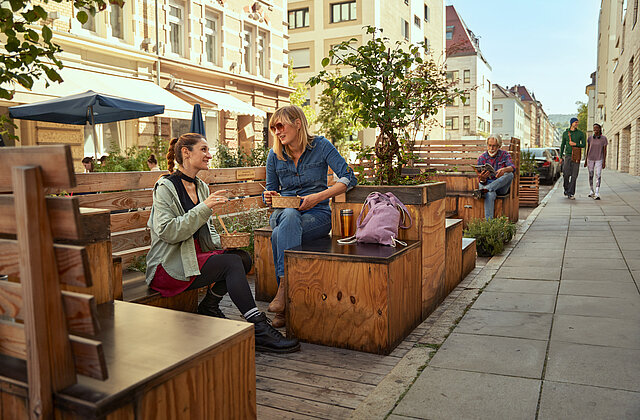 Foto: Zwei Frauen sitzen in einem Parklett (Bänke und Pflanzen, die auf einem Parkplatz am Straßenrand aufgestellt wurden)