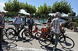 Gruppe von fünf Personen mit Fahrrädern auf einem Weg am Wasser unter Bäumen bei sonnigem Wetter