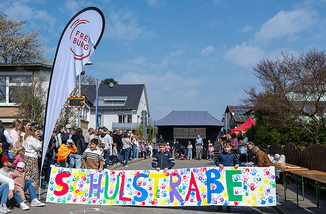 Kinder spannen ein buntes Banner, auf dem Schulstraße steht, quer über eine Straße. Rechts und links stehen weitere Kinder und Erwachsene am Straßenrand