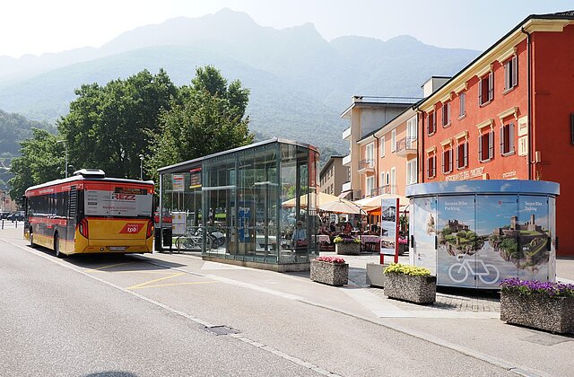 Stadtbus an einer Haltestelle mit Glaswartehäuschen und Fahrradabstellanlage vor mehrstöckigen Gebäuden im Hintergrund vor Bergkulisse