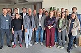 Gruppe von Menschen in einem hellen Raum, einige stehen in lockerer Reihe, eine Frau in der vorderen Reihe trägt eine gemusterte Bluse und eine rote Hose