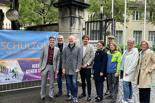Acht Personen stehen seitlich neben einem zwischen zwei Torpfosten gespannten Banner, auf dem Schulzone steht.