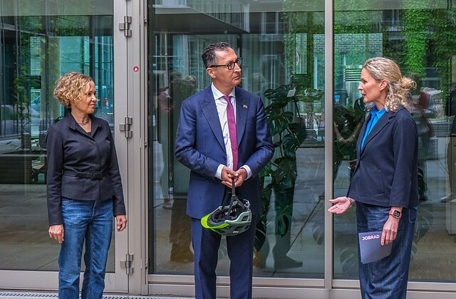 Drei Personen stehen vor einem Glasgebäude, die mittlere Person hält einen Fahrradhelm in der Hand.