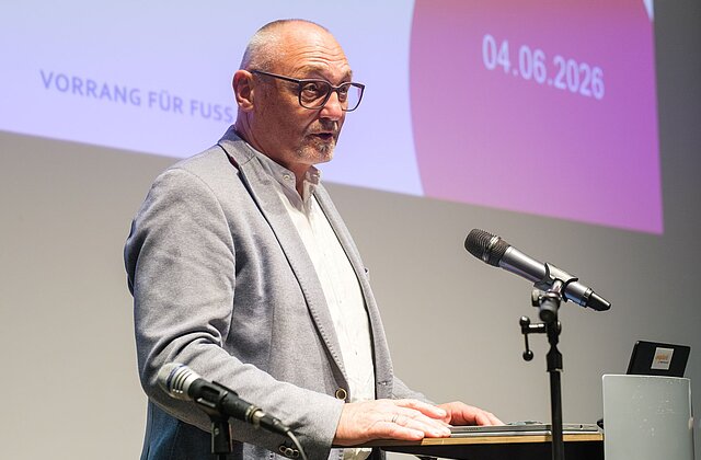 Mann mit Brille und grauem Sakko spricht an einem Rednerpult mit Mikrofonen vor einer Projektionswand