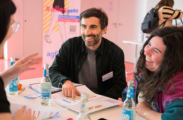 Drei Personen sitzen an einem Tisch mit Getränkeflaschen und Unterlagen, ein Mann zeigt auf ein Blatt und alle lachen oder lächeln.
