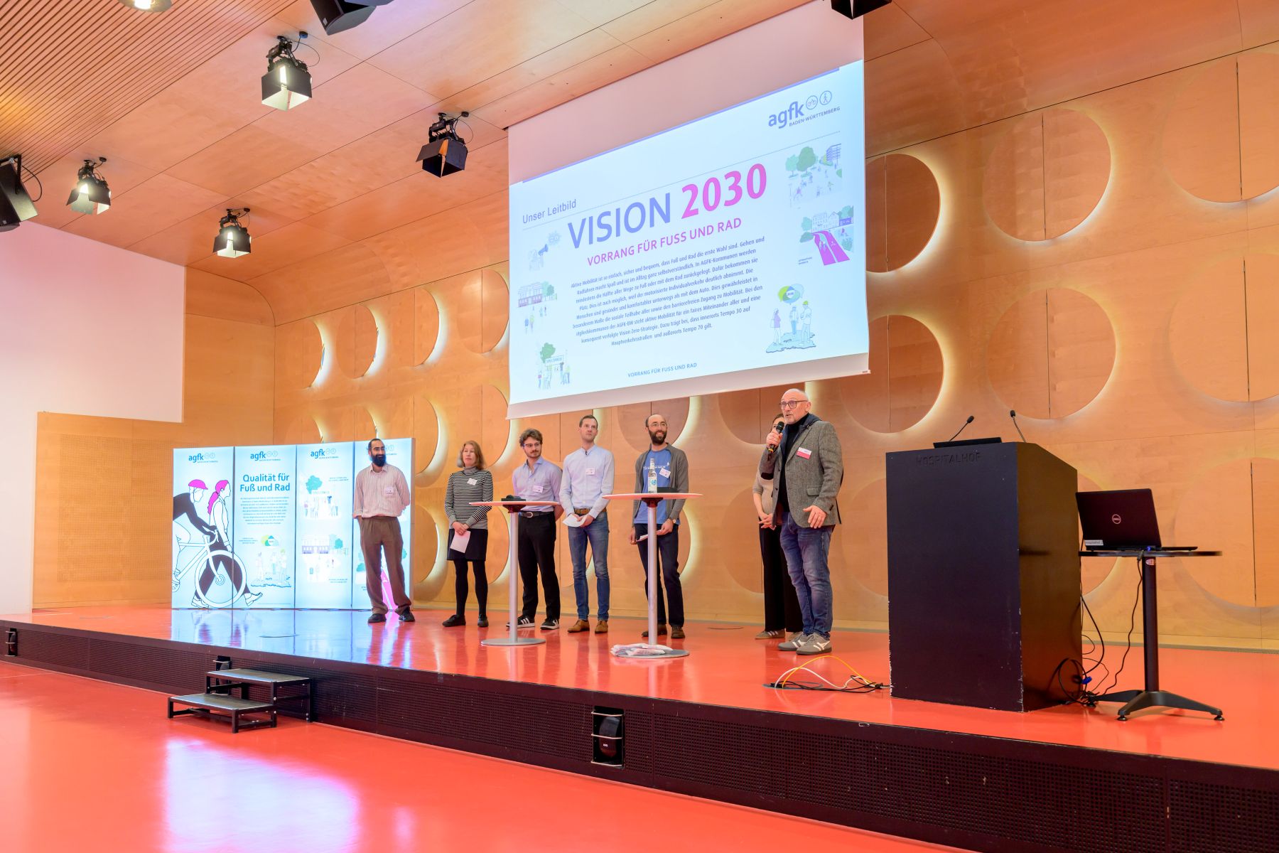 Sieben Personen stehen auf einer Bühne vor einer großen Leinwand mit der Überschrift 'VISION 2030' und mehreren Roll-ups mit Illustrationen und Texten im Hintergrund