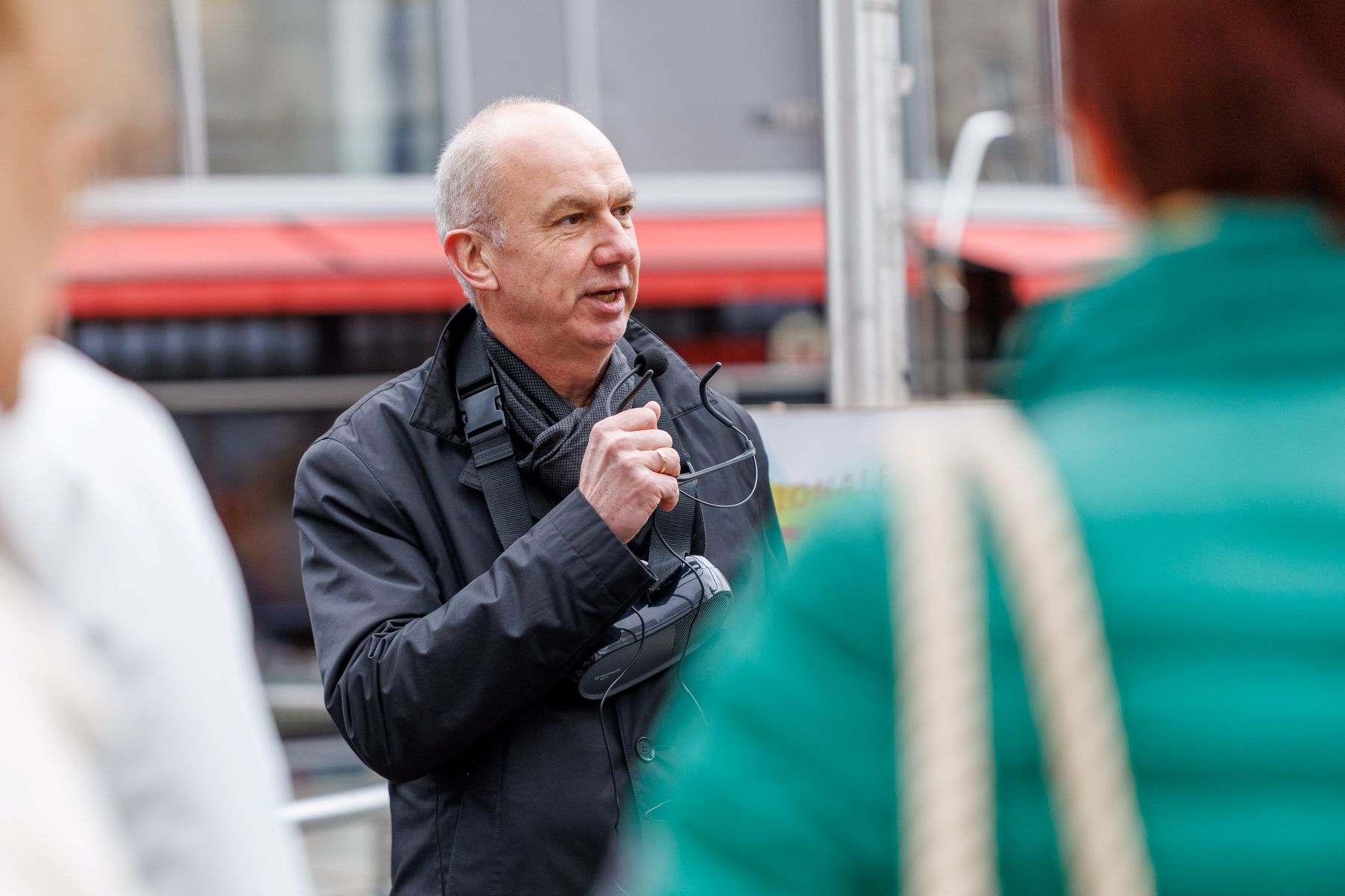 Älterer Mann mit grauen Haaren und schwarzer Jacke spricht in ein Mikrofon vor einer Gruppe von Menschen im Freien