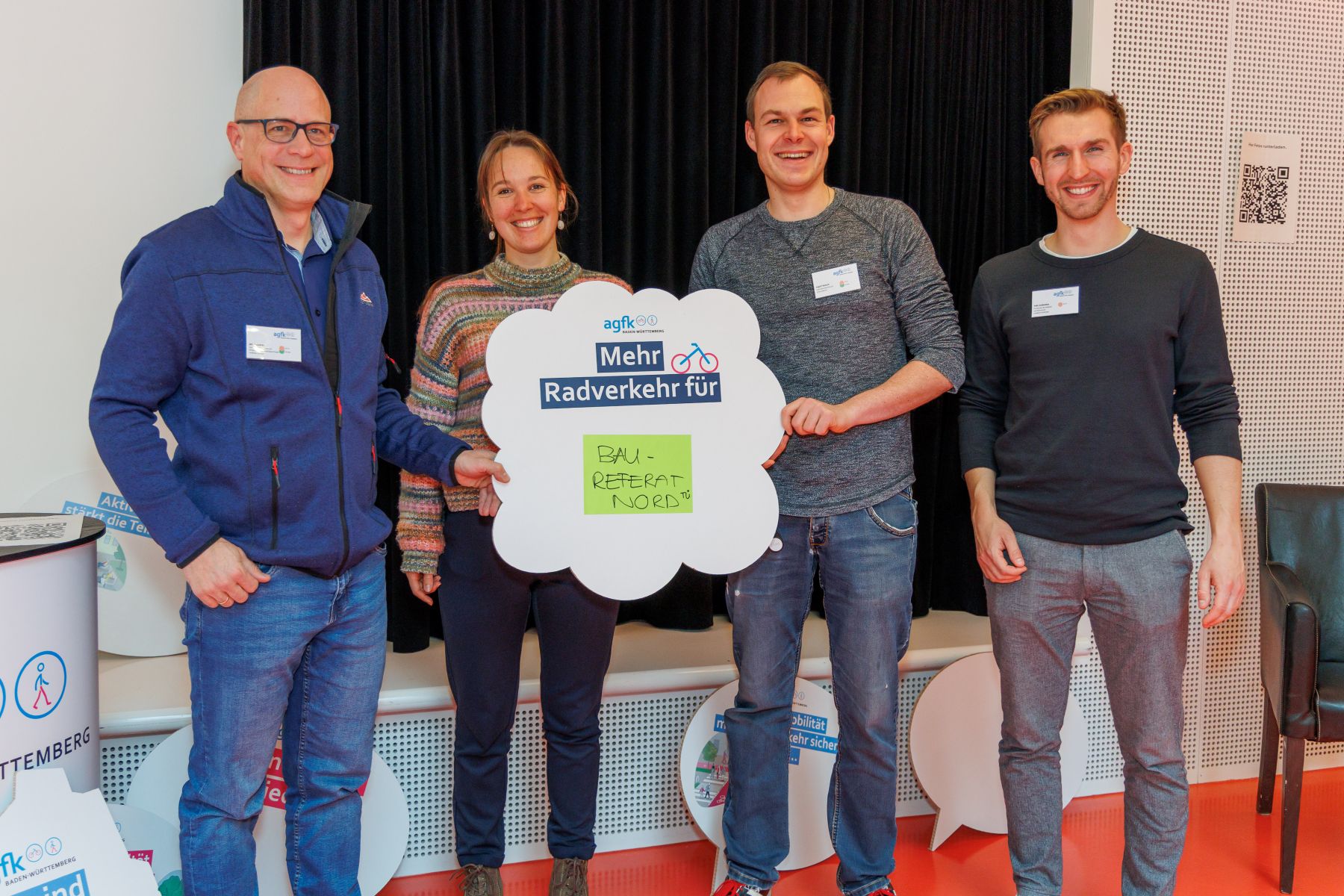 Vier Personen stehen nebeneinander und halten ein schildförmiges Plakat mit dem Text 'Mehr Radverkehr für' und einem gelben Zettel mit der Aufschrift 'BAUREFERAT NORD'
