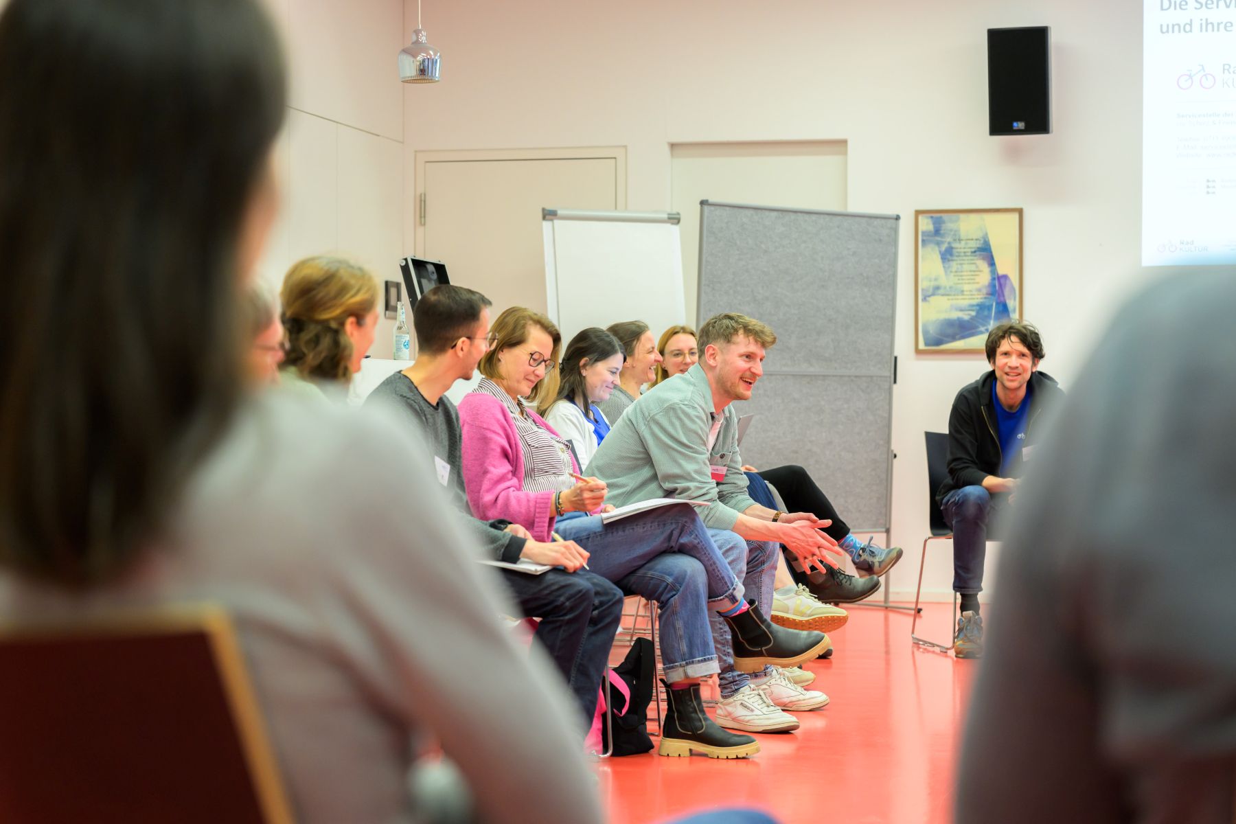 Gruppe von Teilnehmenden sitzt in einem Stuhlkreis in einem hellen Raum mit rotem Boden und diskutiert