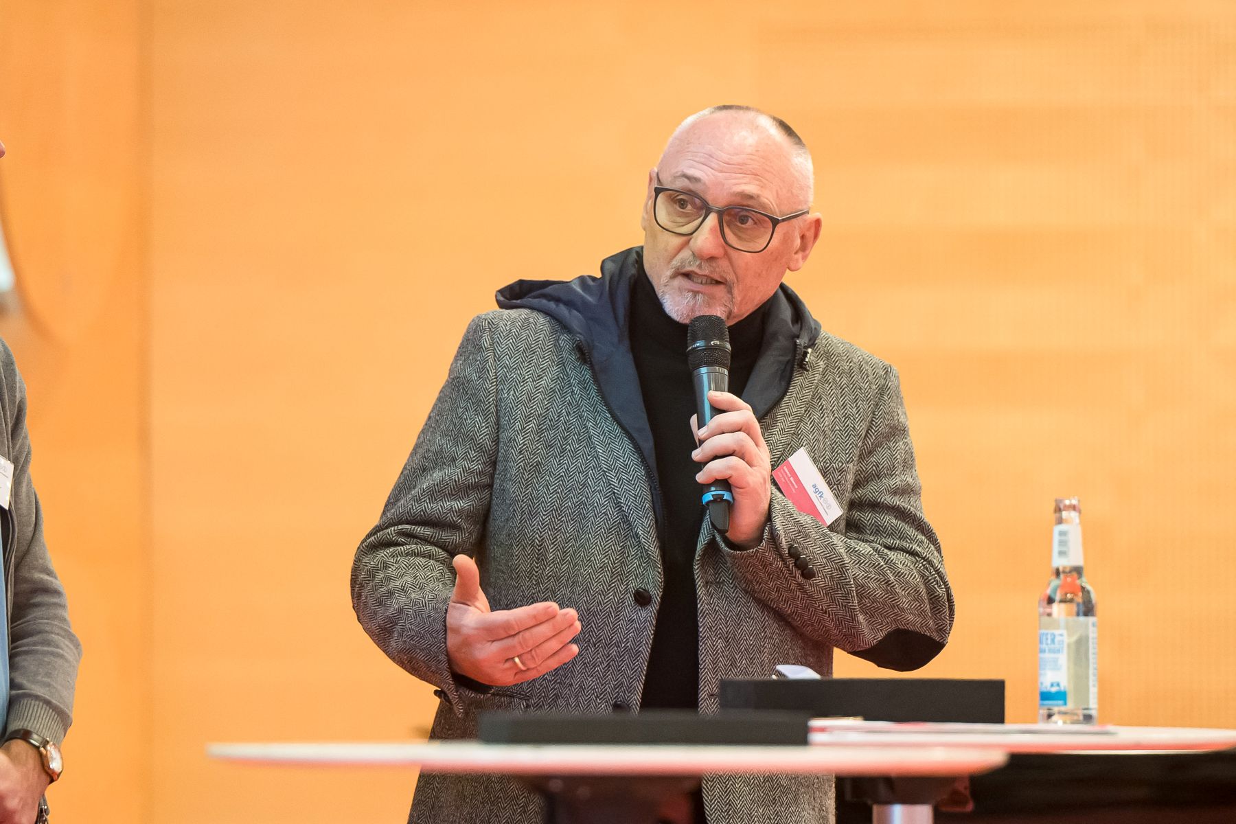 Mann mit grauem Sakko (AGFK-BW-Vorsitzender Günther Riemer) hält Mikrofon und gestikuliert vor einem Tisch mit Wasserflasche und Unterlagen