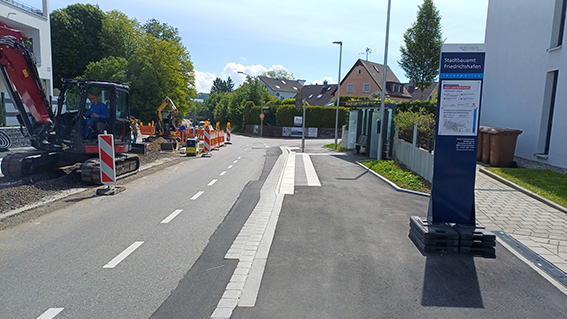 Eine Infostele steht am Rand einer innerörtlichen Straße, an der gebaut wird