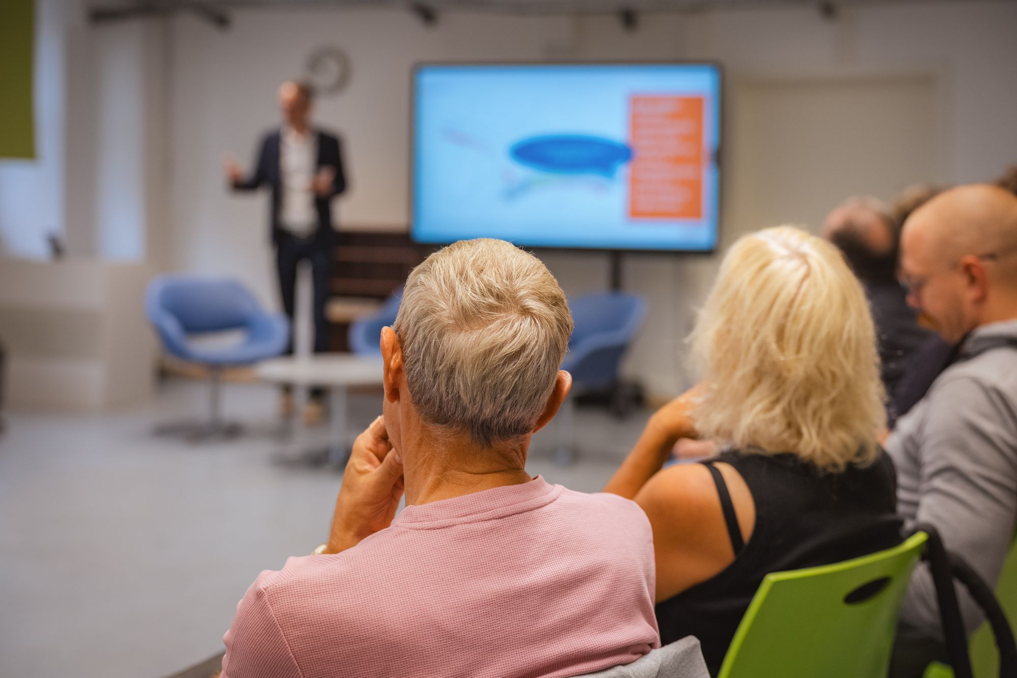 Rückansicht von drei sitzenden Personen, die einem stehenden Vortragenden mit Präsentation auf einem Bildschirm folgen
