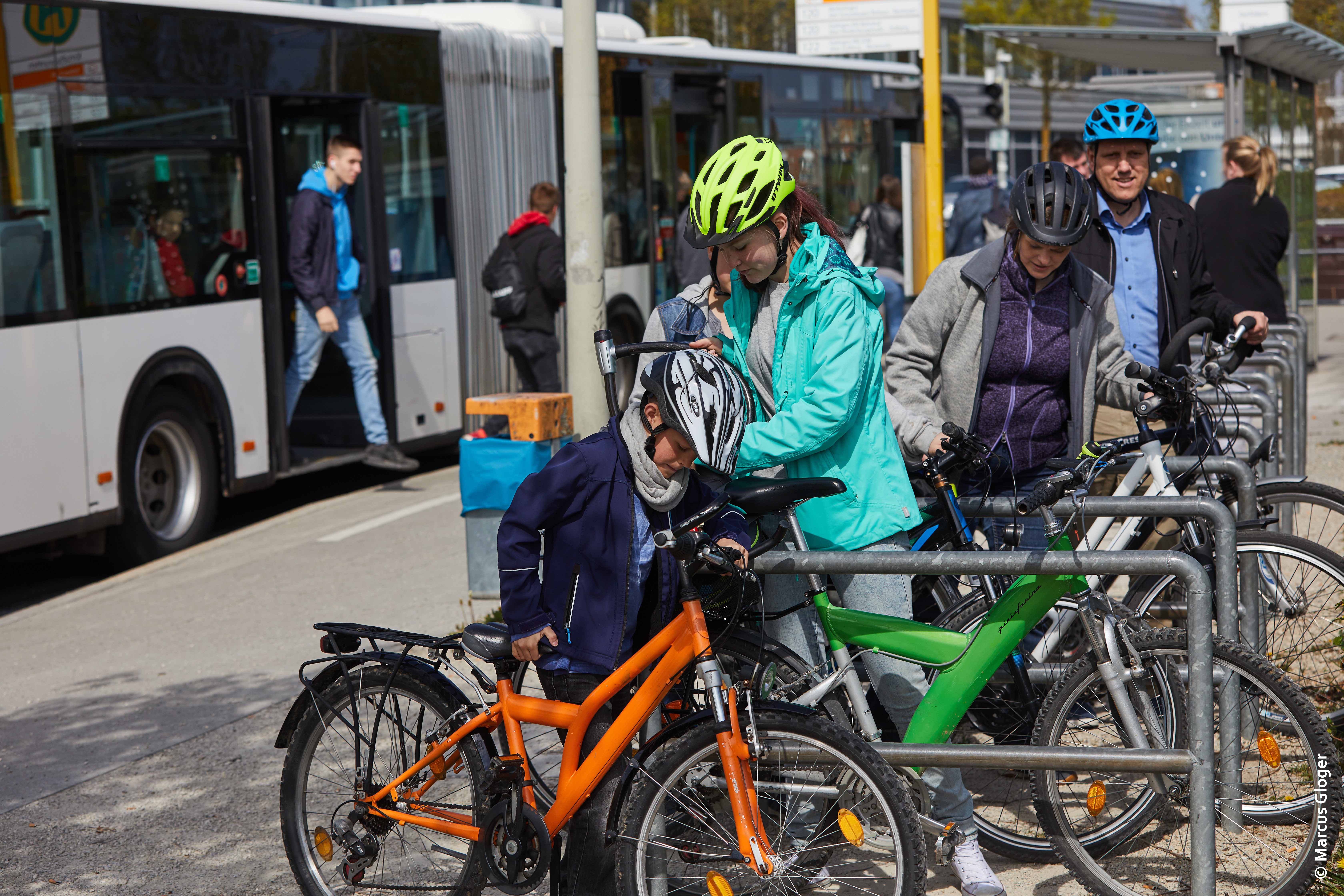Mehrere Personen mit Fahrrädern stehen an einem Fahrradständer in der Nähe einer Bushaltestelle. Eine Person in einer türkisfarbenen Jacke hilft einem Kind, das sein Fahrrad abstellt. Im Hintergrund steigen Personen aus einem Bus.