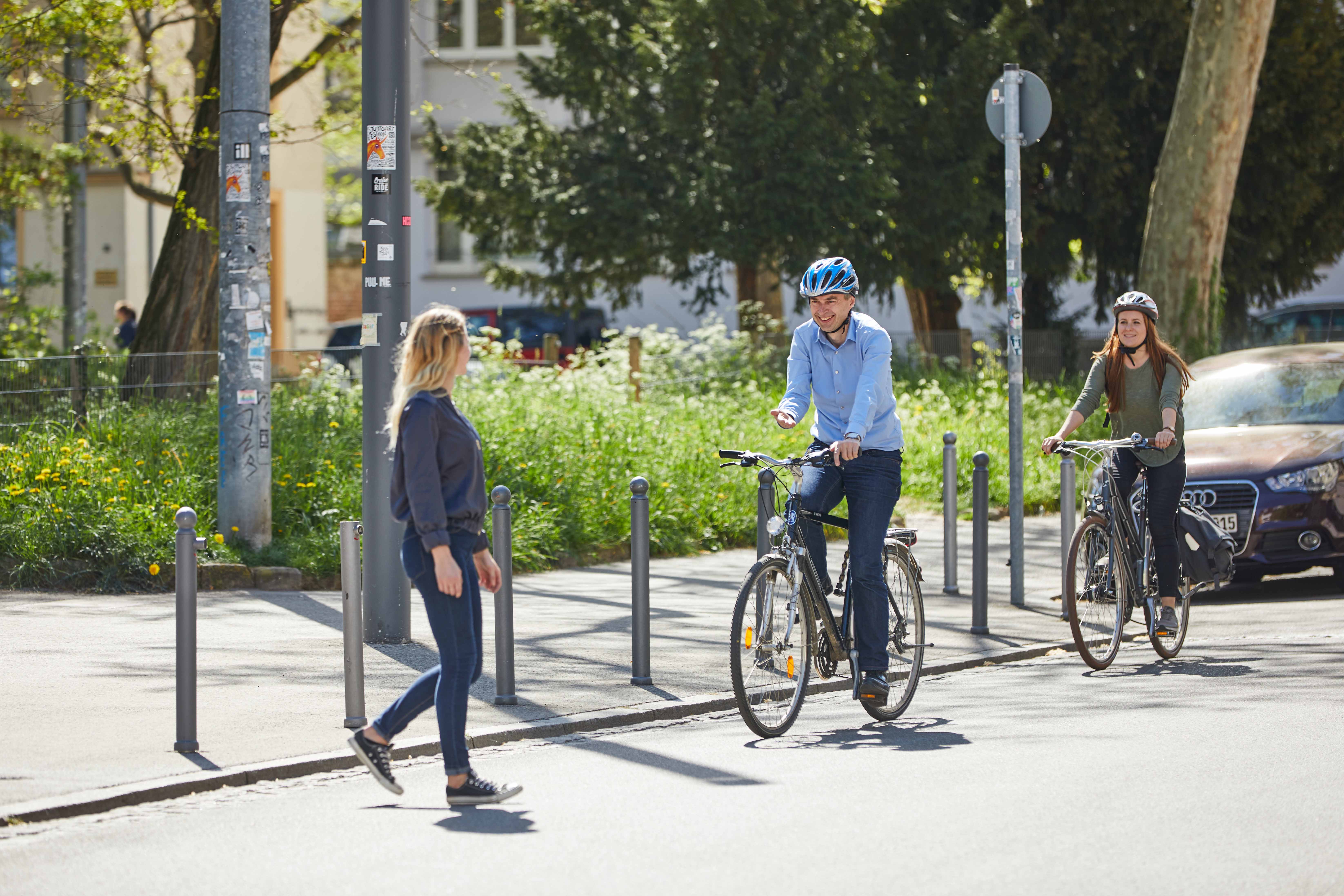 Zwei Radfahrende lassen eine Fußgängerin vor, die die Straße überqueren möchte.