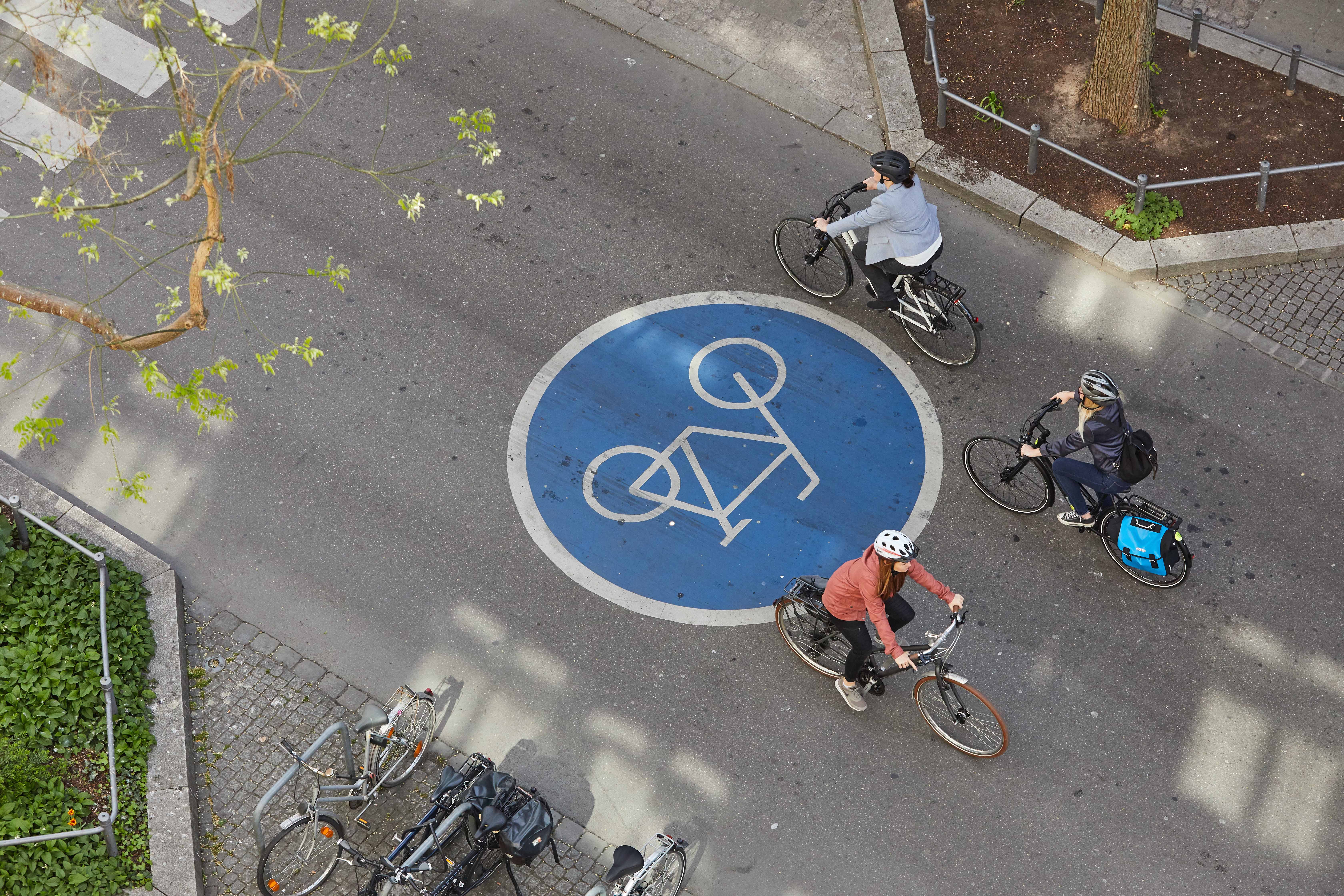 Vier Radfahrende bewegen sich auf einer Fahrbahn mit großem blauen Fahrradsymbol, das eine Fahrradstraße oder eine besondere Radverkehrsführung kennzeichnet. Die Szene wird aus der Vogelperspektive gezeigt, mit umliegendem Gehweg, Begrünung und geparkten Fahrrädern.