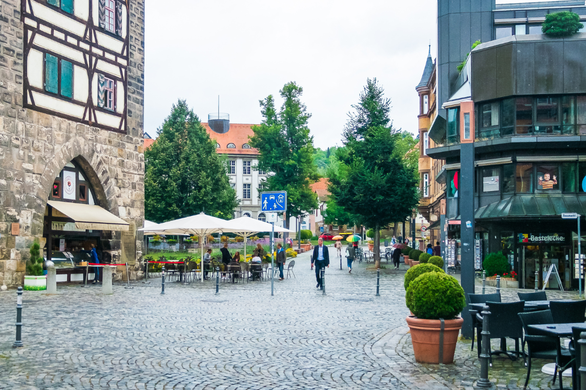 Belebte Fußgängerzone mit Kopfsteinpflaster, Straßencafés unter Sonnenschirmen, Menschen mit Regenschirmen, historischen Fachwerk- und modernen Gebäuden sowie einem blauem Verkehrsschild für den verkehrsberuhigten Bereich.