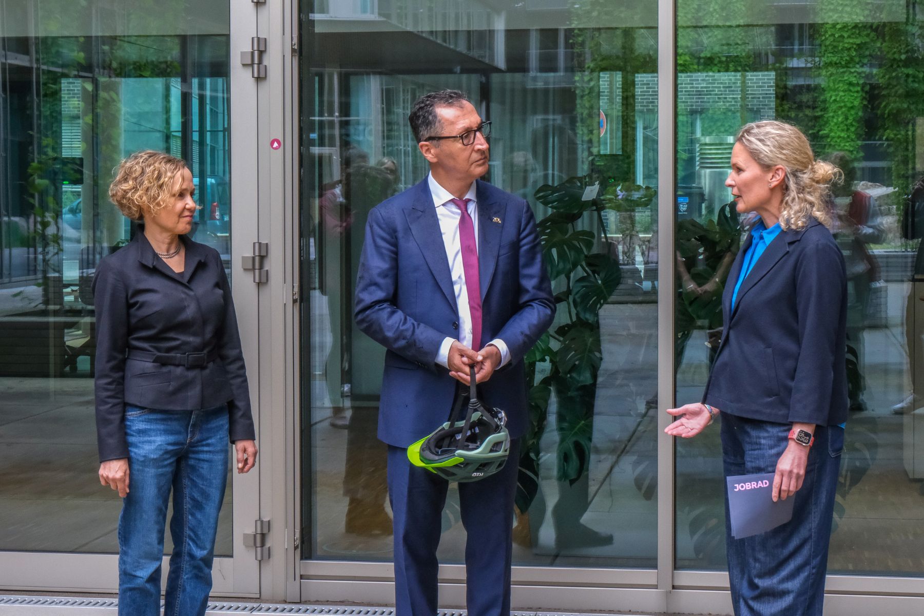 Drei Personen stehen vor einem Glasgebäude, die mittlere Person hält einen Fahrradhelm in der Hand.