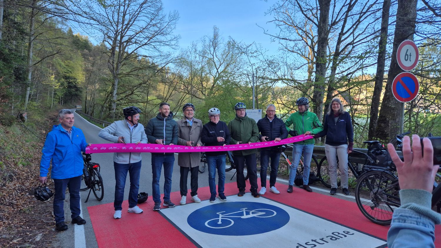 Gruppe von Menschen mit Fahrradhelmen steht auf roter Fahrradstraße mit blauem Fahrradsymbol, eine Person hält ein langes pinkfarbenes Band, im Waldgebiet mit Bäumen im Hintergrund