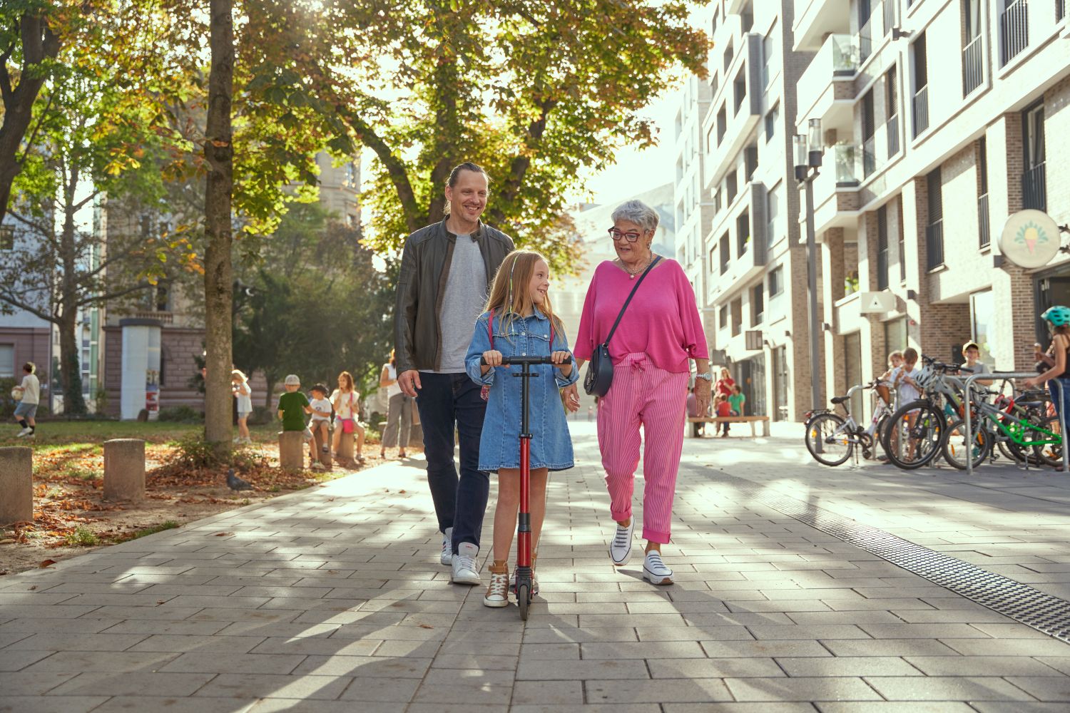 Vater, Tochter und Oma spazieren durch ein Wohnquartier. Die Tochter fährt auf einem Tretroller.