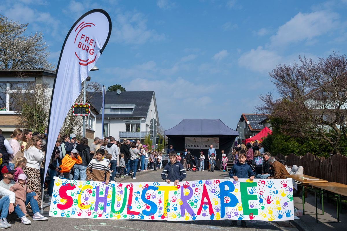 Kinder spannen ein buntes Banner, auf dem Schulstraße steht, quer über eine Straße. Rechts und links stehen weitere Kinder und Erwachsene am Straßenrand
