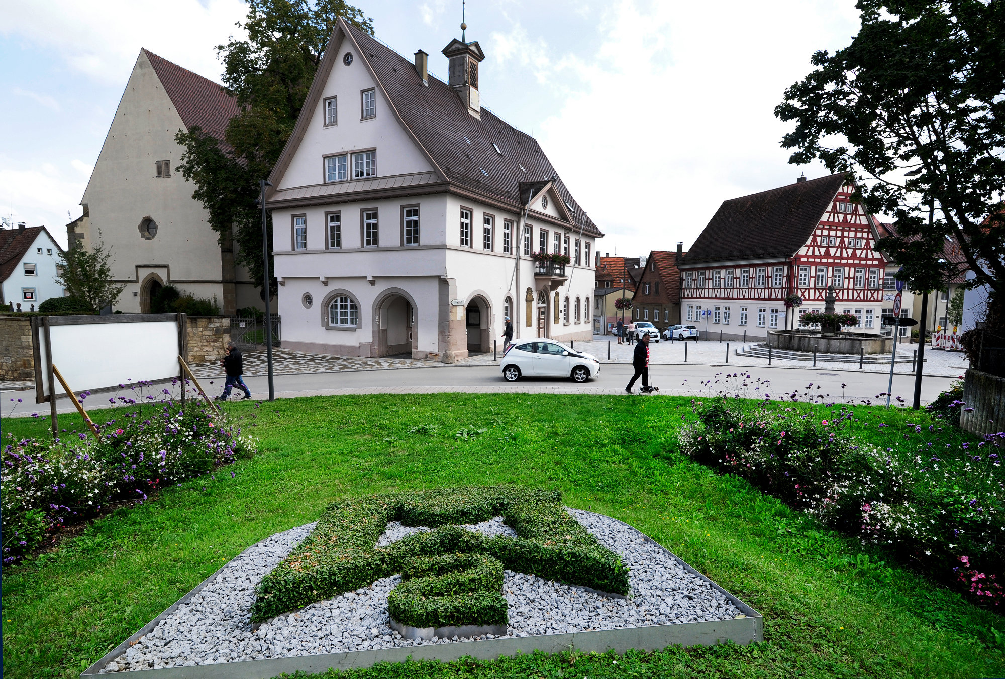 Blick auf einen grünen Platz mit einem Heckenornament in Form eines Wappens, umgeben von Blumenbeeten, davor historische Fachwerkhäuser und ein weißes Auto auf der Straße