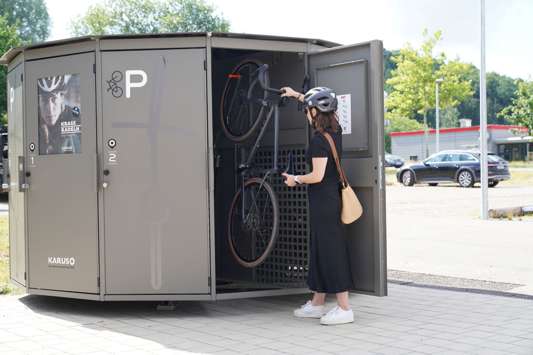 Frau mit Fahrradhelm stellt Fahrrad in einen grauen Fahrradparkcontainer mit offener Tür auf einem Bürgersteig
